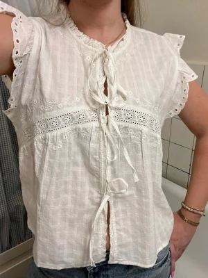Vit blus med knyt och spetsdetaljer - Söt vit blus från Indiska med broderade spetsdetaljer och tre knytband framtill. Blusen har korta ärmar med volang och hålmönster, samt rundad halsringning. Tillverkad i ett lätt och luftigt tyg, perfekt för sommaren.