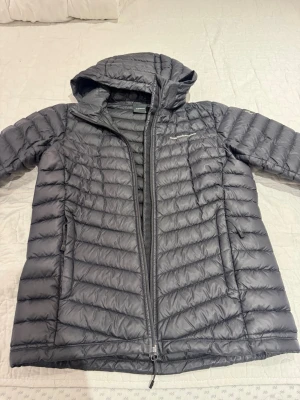 Svart dunjacka Peak Performance - Snygg svart dunjacka från Peak Performance med huva och dragkedja framtill. Jackan har två sidofickor med dragkedja och diskret logga på bröstet. Lättviktigt material och quiltad design gör den perfekt för kyliga dagar.