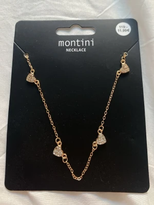 Guldfärgat halsband med hjärtan Montini - Snyggt guldfärgat halsband från Montini med små hjärtformade hängen dekorerade med glittrande pärlor. Kedjan är tunn och elegant, perfekt för att ge en söt touch till din outfit. Hjärtana är jämnt placerade längs kedjan för en balanserad look.
