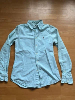 Ljusblå skjorta från Ralph Lauren - Klassisk ljusblå skjorta från Ralph Lauren med broderad logga på bröstet. Skjortan har lång ärm, knappar framtill och krage. Perfekt för en stilren look och enkel att matcha med jeans eller chinos. Priset ej hugget i en sten 