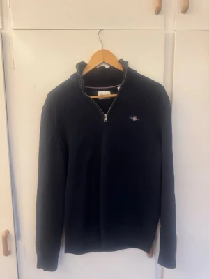 Mörkblå stickad tröja från Gant - Snygg mörkblå stickad tröja från Gant med halv dragkedja och liten broderad logga på bröstet. Tröjan har hög krage och långa ärmar, perfekt för lager på lager. Klassisk och stilren design som passar till det mesta. Ny kostar 1,200kr ca