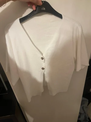 Vit kortärmad kofta med knappar - Snygg vit kofta med korta ärmar och två dekorativa knappar framtill. Koftan har en djup v-ringning och är tillverkad i ett tunt, mjukt material som passar perfekt till sommaren. Enkel och stilren design som är lätt att matcha.