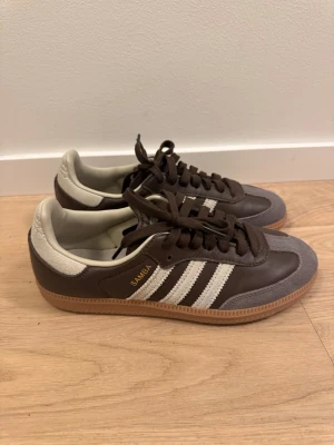 Adidas Samba bruna sneakers - Säljer ett par klassiska Adidas Samba sneakers i brunt läder med grå mockadetaljer och vita ränder på sidorna. Gummisula och guldfärgad SAMBA-text. De är inköpta i London men är tyvärr lite för små, därför måste jag tyvärr sälja dem.