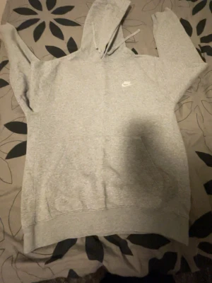 Grå hoodie från Nike med logga - Säljer en klassisk grå hoodie från Nike med vit broderad logga på bröstet. Hoodien har huva med dragsko och en stor magficka framtill. Tillverkad i mjukt bomullsmaterial som är perfekt för chill dagar eller streetwear-stil.