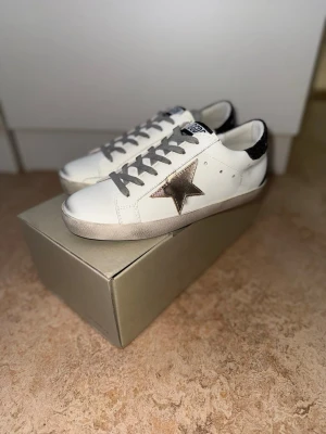 Vita golden goose  - Snygga vita sneakers från Golden Goose med metallicfärgad stjärna på sidan och svarta glittriga detaljer vid hälen. Skorna har grå skosnören och en klassisk låg siluett. Tillverkade i läder med slitstark gummisula. Perfekta för en trendig och avslappnad stil.