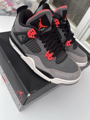 Air Jordan 4 Retro grå/röd sneakers - Snygga Air Jordan 4 Retro sneakers i grå mocka med svarta och röda detaljer. Skorna har svart mesh på sidorna, svarta skosnören och en vit/grå sula. Ikonisk Jumpman-logga på plösen och hälen. Perfekta för dig som vill sticka ut med stilrena och sportiga sneakers.