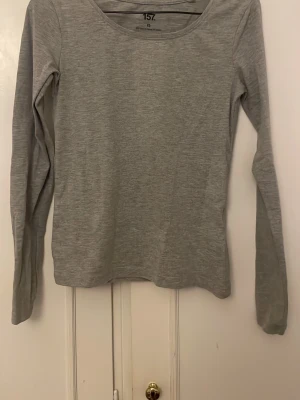 Ljusgrå långärmad topp från 157 - En enkel och stilren ljusgrå långärmad topp från 157. Toppen har rund halsringning och är tillverkad i mjukt material som känns skönt mot huden. Perfekt att matcha med jeans eller kjol för en clean look.