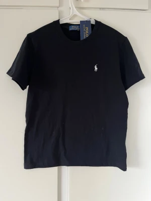 Svart t-shirt från Polo Ralph Lauren - Klassisk svart t-shirt från Polo Ralph Lauren med rund hals och korta ärmar. T-shirten har den ikoniska vita logobroderingen på bröstet och är tillverkad i mjuk bomull för en skön känsla. Perfekt till jeans eller shorts.