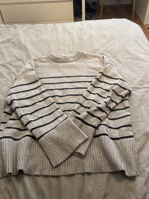 Randig stickad tröja  - Grå stickad tröja med svarta horisontella ränder från 157. Tröjan har rund halsringning, långa ärmar och breda ribbade muddar vid ärmslut och nederkant. Perfekt för en avslappnad och stilren look.