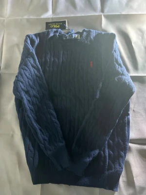 Mörkblå kabelstickad tröja Polo Ralph Lauren - Kabelstickad mörkblå tröja från Polo Ralph Lauren med klassisk röd logga broderad på bröstet. Tröjan har rund halsringning, långa ärmar och ribbade muddar. Skriv vid funderingar. Pris kan diskuteras vid snabb affär.