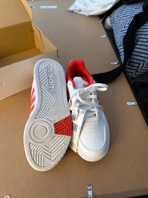 Vita och röda Adidas sneakers - Säljer snygga vita sneakers från Adidas med klassiska röda ränder på sidorna och röd insida. Skorna säljs för att dom inte används. Skorna har aldrig används, dom är helt nya. Priset går att diskuteras.