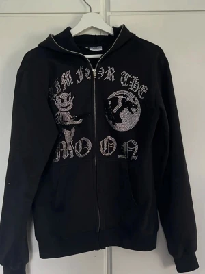 Svart hoodie med tryck och dragkedja - Snygg svart hoodie med dragkedja och huva. Framsidan har ett stort tryck med texten 'Aim for the moon' och motiv av en tecknad figur och en måne i glittrande stenar. Perfekt för dig som gillar streetwear och coola detaljer.