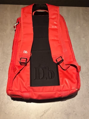 Röd ryggsäck från Db -  Röd ryggsäck från Db med svarta detaljer och broderad Db-logga på ryggen. Ryggsäcken har justerbara axelband och är tillverkad i slitstarkt syntetmaterial. Fint skick.