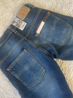 Mörkblå nudie jeans slim fit - Säljer ett par mörkblå jeans med raka ben och diskreta slitningar på låren. Jeansen har klassisk femficksdesign och kontrastsömmar. Perfekta för en avslappnad och stilren look. Slim fit 