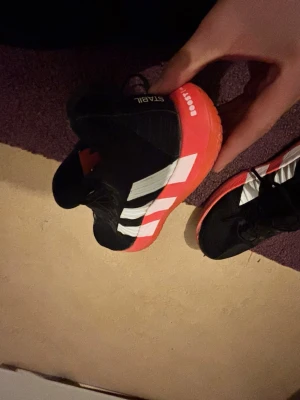 Adidas svart-röda träningsskor - Säljer ett par snygga Adidas träningsskor med svart ovandel i mesh och syntet, vita klassiska ränder på sidan och röd sula med BOOST-dämpning. Skorna har snörning och låg profil, perfekta för inomhussport, gym eller Handbollskor