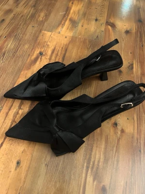 Svarta pumps med rosett - Stilrena svarta pumps med spetsig tå och dekorativ rosett framtill. Skorna har öppen häl med justerbart spänne och är tillverkade i ett satinliknande material. Klacken är 5cm. Aldrig använda 