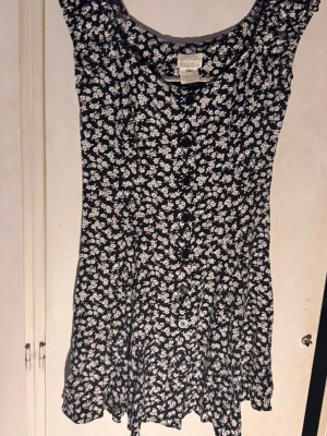 Svart blommig vintageklänning med knappar - Supersöt svart vintage kortklänning med vitt blommönster och knappar framtill. Klänningen har en lätt utställd passform och är ärmlös, perfekt för varma dagar. Tillverkad i mjuk och skön viskoskvalitet. 100% viskos (rayon)