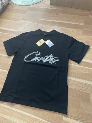 Black Corteiz T-Shirt - Snygg svart t-shirt från Critiz med ett stort, silvrigt graffiti-inspirerat tryck på bröstet. Klassisk passform med korta ärmar och rund hals. Tillverkad i mjuk bomull som känns skön mot huden. Perfekt för dig som gillar streetwear och vill sticka ut.