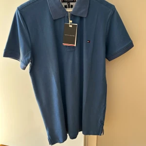 Blå pikétröja från Tommy Hilfiger - Snygg blå pikétröja från Tommy Hilfiger med klassisk krage och liten logga på bröstet. Tillverkad i mjuk bomull och har korta ärmar. Perfekt till jeans eller shorts för en clean look. Helt oanvänd.