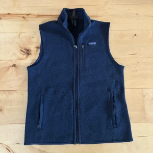 Marinblå fleeceväst från Patagonia - Säljer en marinblå fleeceväst från Patagonia med hög krage och dragkedja framtill. Den köptes förra året från naturkompaniet för 1400kr men passade inte riktigt mig. Den är tvättad en gång vilket resulterade i lite noppror som går att få bort med en knoppifier. Västen har tre fickor med dragkedja och en klassisk logga på bröstet. Perfekt lager för kyliga dagar och enkel att matcha med olika outfits.
