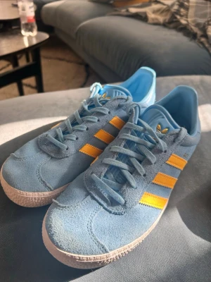 Adidas Gazelle blå/gula sneakers 38 - Snygga Adidas Gazelle sneakers i ljusblå mocka med klassiska gula ränder på sidorna och vit sula. Skorna har lågt skaft och blå snörning. Knappast andvända storlek 38 