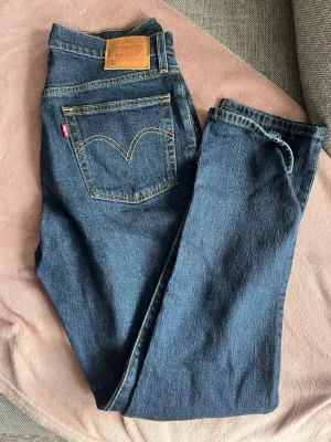 Levi's blå raka jeans - Klassiska blå jeans från Levi's med rak passform och hög midja. Jeansen har fem fickor, knappgylf och den ikoniska röda Levi's-lappen på bakfickan. Tillverkade i slitstark denim med kontrastsömmar. Perfekta för en avslappnad och tidlös stil.