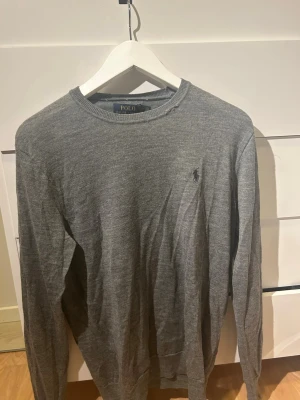 Grå merinoulltröja Polo Ralph Lauren - Slim fit grå tröja från Polo Ralph Lauren i mjuk och tvättbar merinoull. Klassisk rund hals och diskret broderad logga på bröstet. Långärmad och stilren, perfekt för lager-på-lager under höst och vinter. Liten defekt vid halsen.