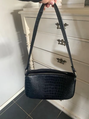 Svart axelväska i crocopräglat skinn - Snygg svart handväska med crocopräglat mönster och axelrem. Väskan har en stilren, rektangulär form och dragkedja upptill. Perfekt för dig som gillar trendiga accessoarer med lite edge.