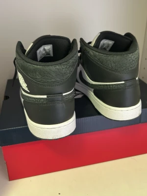 Air Jordan 1 Mid svart/vit - Säljer ett par Air Jordan 1 Mid sneakers i svart och vitt läder med klassisk Swoosh och Air Jordan-logga på sidan. Skorna har svart mönstrad detalj runt kragen och vit sula. Perfekta för dig som gillar streetwear och ikonisk design.
