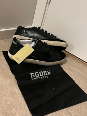 Svarta Golden Goose sneakers med glitter - Stilrena svarta sneakers från Golden Goose med vit sula och svart stjärndetalj på sidan. Hälpartiet är täckt av svart glitter och har logotypen 'Golden Goose' i vitt. Skorna har klassisk låg profil och är tillverkade i läder.