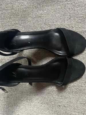 Svarta pumps med öppen tå från NLY - Stilrena svarta pumps från NLY med öppen tå och smal rem över foten. Skorna har justerbart spänne vid ankeln och en klassisk, elegant design. Materialet ser ut att vara konstläder eller mockaimitation. Perfekta för att lyfta vilken outfit som helst.