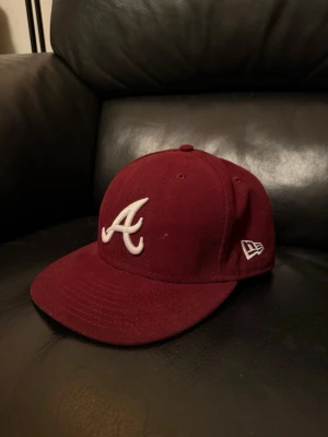 Vinröd New Era Atlanta keps, storlek 7 3/8 - Säljer en vinröd keps från New Era med Atlanta Braves-logga broderad i vitt framtill och New Era-logga på sidan. Klassisk rak skärm och justerbar passform. Tillverkad i bomullstyg som ger en skön känsla och snygg look.