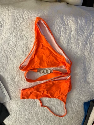Orange asymmetrisk baddräkt - Säljer en snygg orange baddräkt med asymmetrisk design och en axelband. Baddräkten har dekorativa ringar i metall i midjan som ger en unik look. Materialet är stretchigt och snabbtorkande, perfekt för stranden eller poolen.