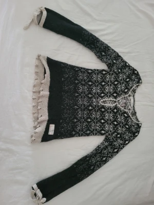 Svartvit stickad tröja med mönster - Säljer en svartvit stickad tröja med snyggt nordiskt mönster. Koftan har långa ärmar, knytband i midjan och dekorativa band vid ärmsluten. Fina beige detaljer längs kanterna och volangkant nedtill ger extra stilpoäng. Perfekt för dig som gillar unik stickad design.