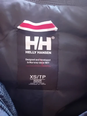 Mörkblå parkas Helly Hansen XS - Snygg mörkblå parkas från Helly Hansen i storlek XS. Jackan har huva med fejkpäls och är designad för att hålla dig varm under kalla dagar. Klassisk modell med regular fit och tåligt yttertyg. Perfekt för dig som vill ha både stil och funktion.