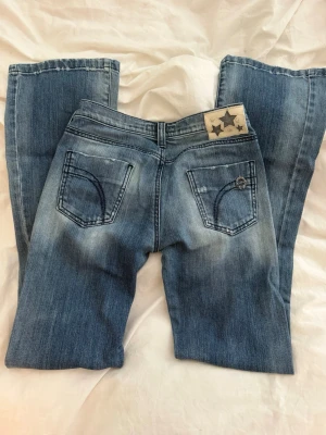 Killah low rise jeans  - Säljer ett par blå bootcut jeans från Killah med snygg slitning och dekorativa bakfickor. Jeansen har låg midja och ett coolt patch med stjärnor bak i linningen. De är storlek 26, passar nog de som brukar använda s i byxor 
