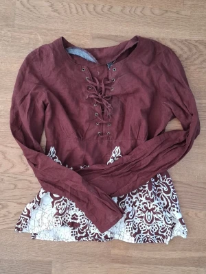 Vinröd topp med snörning och vitt mönster - Unik vinröd topp med lång ärm och snörning framtill. Nedre delen har ett dekorativt vitt mönster. I gott skick. 