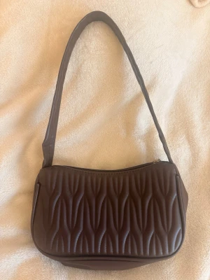 Brun  axelremsväska🤎🥥 - Snygg brun handväska med quiltat mönster och mjuk struktur. Väskan har en bred axelrem och stängs med dragkedja upptill. Perfekt för dig som gillar trendiga accessoarer med unik design. Materialet ser ut att vara konstläder.
