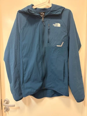 Blå vindjacka från The North Face - Snygg blå vindjacka från The North Face med huva och hel dragkedja. Jackan har ett diskret rutmönster, bröstficka med dragkedja och logga på vänster sida. Tillverkad i lätt och slitstarkt syntetmaterial, perfekt för blåsiga dagar. (Lite dåligt ljus på bilderna)