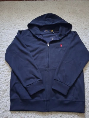 Marinblå hoodie från Polo Ralph Lauren - Snygg marinblå hoodie från Polo Ralph Lauren med dragkedja framtill och klassisk röd logga broderad på bröstet. Tillverkad i mjukt bomullsmaterial och har en rymlig huva. Perfekt för en avslappnad och stilren look.