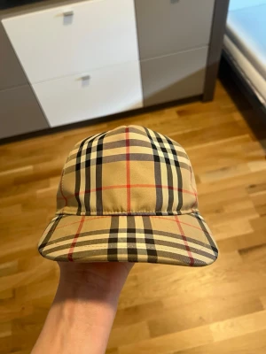 Beige keps med Burberry-mönster - Säljer en beige keps från Burberry med klassiskt rutigt mönster i svart, vitt och rött. Kepsen har en böjd skärm och justerbar rem baktill. Materialet är troligtvis bomull eller polyester och designen är riktigt ikonisk och trendig.