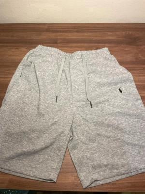 Grå mjukisshorts från Polo Ralph Lauren - Snygga grå mjukisshorts från Polo Ralph Lauren med dragsko i midjan och klassisk logga broderad på vänster ben. Tillverkade i mjukt bomullsmaterial och har en relaxed passform med sidofickor. Perfekta för chill dagar eller träning. Storlek S