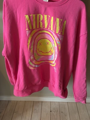 Rosa Nirvana hoodie med tryck - Cool rosa hoodie med stort Nirvana-tryck i gult och vitt på framsidan samt det klassiska smiley-ansiktet. Tröjan har rund halsringning, ribbade muddar och är gjord i mjukt bomullsmaterial. Perfekt för dig som gillar bandmerch och vill sticka ut.
