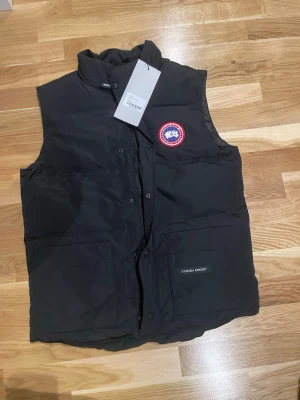 Svart dunväst från Canada Goose - Snygg svart dunväst från Canada Goose med hög krage och två stora framfickor. Västen har ett klassiskt rött och vitt Canada Goose-märke på bröstet och stängs med både dragkedja och knappar. Perfekt för lager-på-lager och streetwear. Storlek S. Skön och perfekt för hela året runt. 