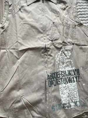 Beige overshirt från Jack & Jones - Stilren beige overshirt från Jack & Jones med dragkedja framtill och tryck med bokstäver och siffror på framsidan. Overshirten har en bröstficka och klassisk krage. Materialet känns som bomull och plagget har en avslappnad streetkänsla.