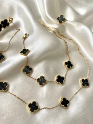 Guldhalsband med svarta klöverhängen - Snyggt halsband i guldton med flera svarta fyrklöverformade hängen. Kedjan är tunn och elegant, och varje hänge har en pärlad kant som ger extra detalj. Perfekt accessoar för dig som gillar stilrena och trendiga smycken.
