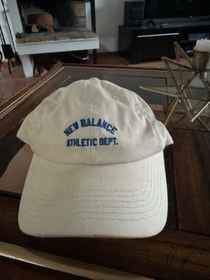 Beige keps från New Balance - Snygg beige keps från New Balance med blå broderad text 'ATHLETIC DEPT.' framtill. Klassisk böjd skärm och justerbart spänne bak. Tillverkad i bomull för en skön och avslappnad look.