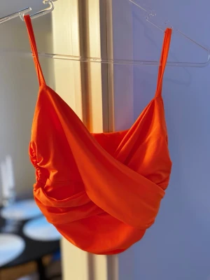 Orange omlott linnetopp - Snygg orange linnetopp med omlottdesign och smala axelband. Toppen har en croppad passform och är tillverkad i ett mjukt, stretchigt material som känns bekvämt mot huden. Perfekt för dig som vill sticka ut med en färgstark look. Endast testad