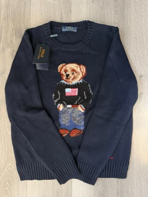 Mörkblå stickad tröja Polo Bear - Mörkblå stickad tröja från Polo Ralph Lauren med ikonisk Polo Bear på bröstet. Björnen har amerikansk flagga på tröjan, jeans och bruna skor. Tröjan har rund halsringning och ribbstickade muddar. Perfekt för dig som gillar klassisk preppy stil.