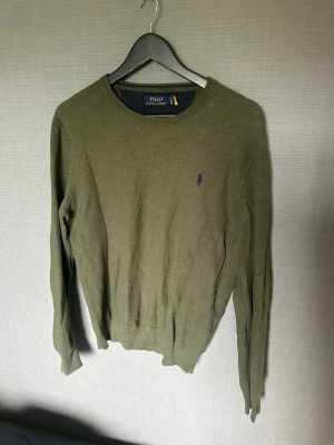 Olivgrön stickad tröja Polo Ralph Lauren - Snygg olivgrön stickad tröja från Polo Ralph Lauren med rund halsringning och diskret mörkblå logga broderad på bröstet. Tröjan har långa ärmar och ribbade muddar vid ärmslut och nederkant. Perfekt för lager på lager och enkel att matcha.